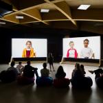 People in a dark exhibition space silhouetted against two screens showing a projection of a film of children talking